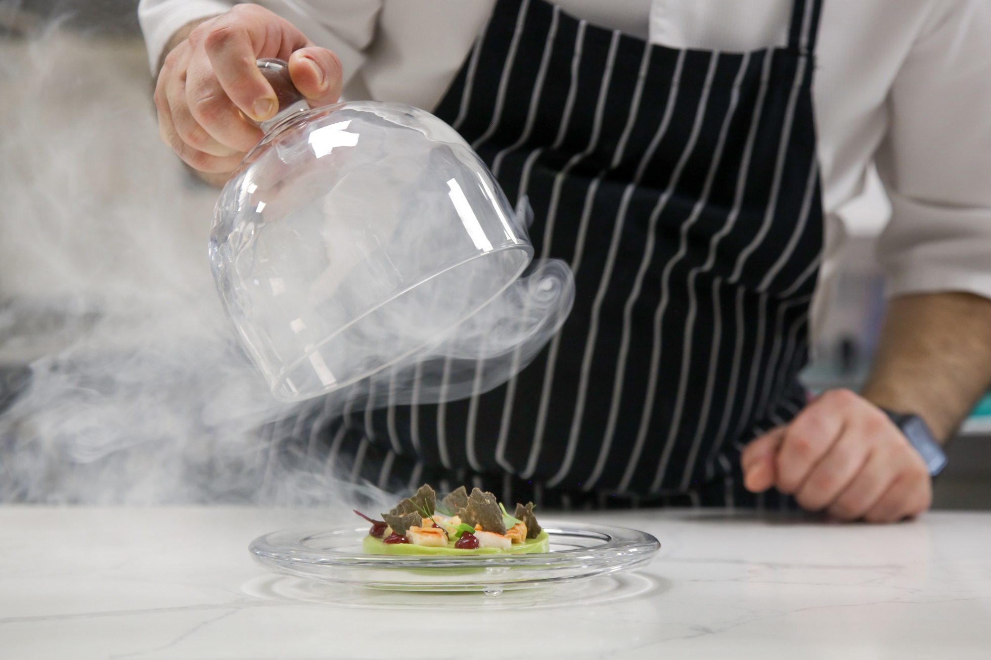 Chef's hand lifts up glass cloche from a plate with hot food and moving smoke at the restaurant